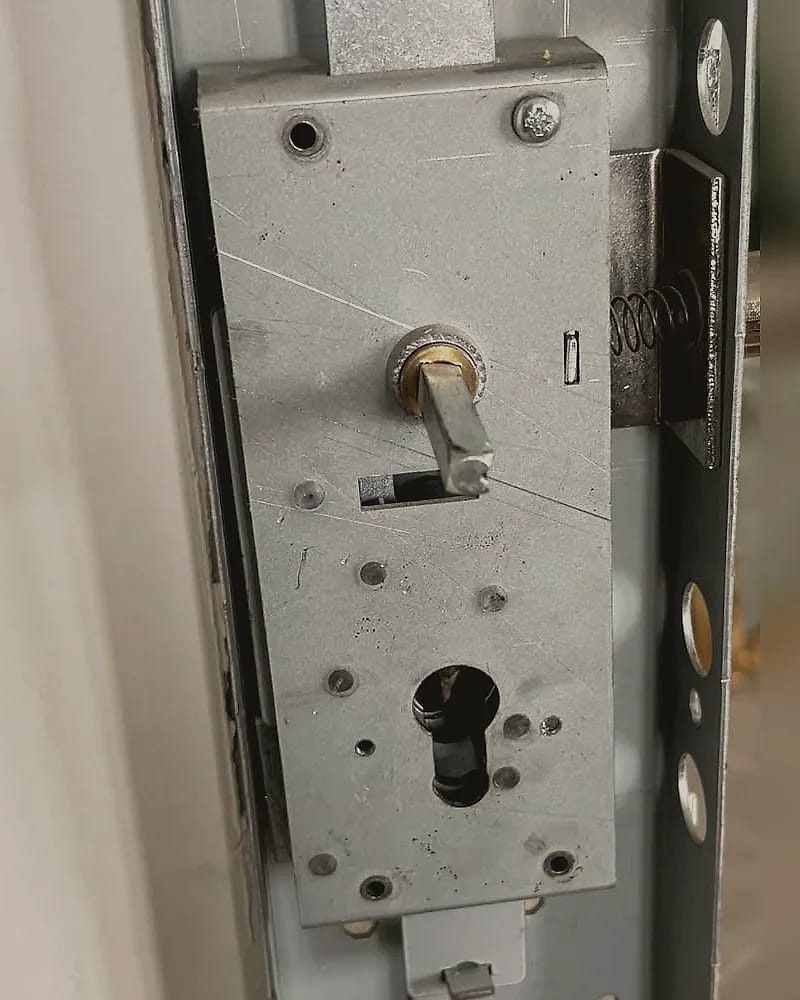 Le boîtier d’une serrure multipoints dans une porte métallique, cylindre retiré : les tringles partent en haut et en bas et la têtière court à droite